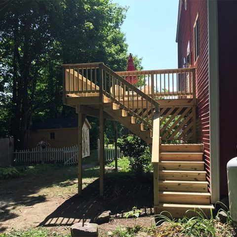 Wooden outdoor deck and stairs beside a red house, with yard and trees in bright sunlight