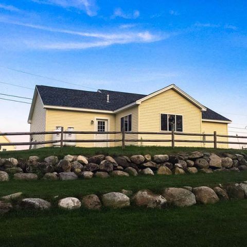 Yellow house with black roof behind a wooden fence, set on a grassy hill with rocks under a blue sky