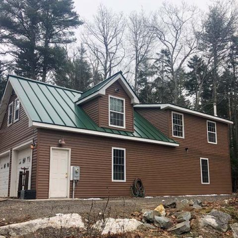 Brown house with green metal roof in a wooded yard, viewed from the front-left.