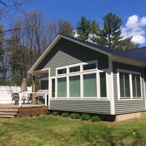 Gray house with white trim, large windows, and a wooden deck in a grassy yard under a blue sky