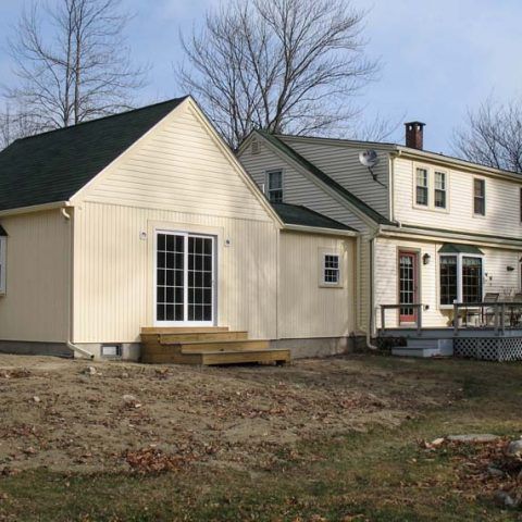 Cream-colored house addition with French doors and a small deck beside a larger home in a bare yard