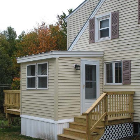 Beige house addition with white trim, front steps, and small deck in a wooded yard.