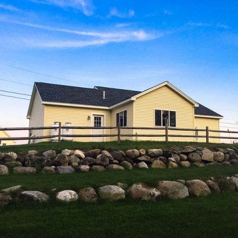 Yellow house with a black roof behind a wooden fence and rock-lined lawn under a blue sky