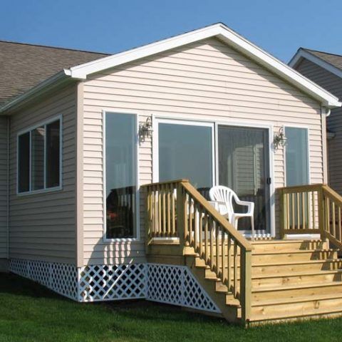 Small beige house porch with wooden steps, railing, and white chairs in a grassy yard
