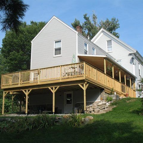 White house with large wooden deck and stairs on a grassy hillside under trees
