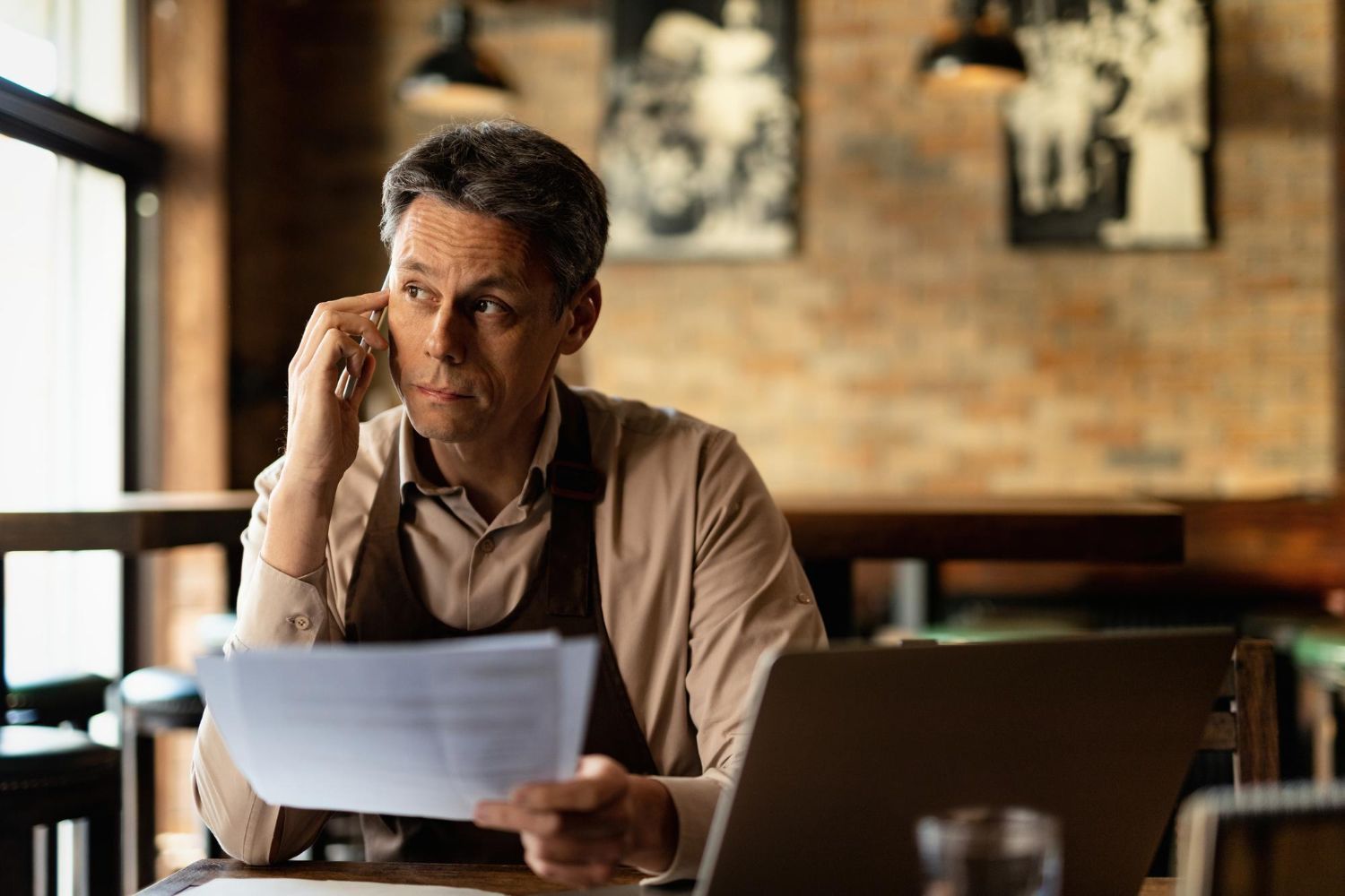 Man reviewing papers at a laptop in a warm café setting, holding one sheet to his ear