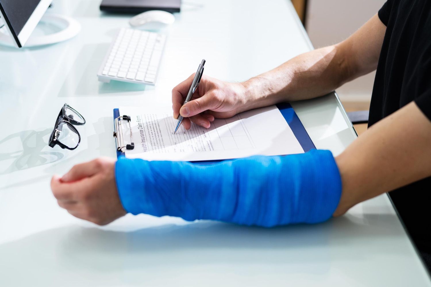 Person with blue cast signing paperwork at a desk beside glasses and a keyboard