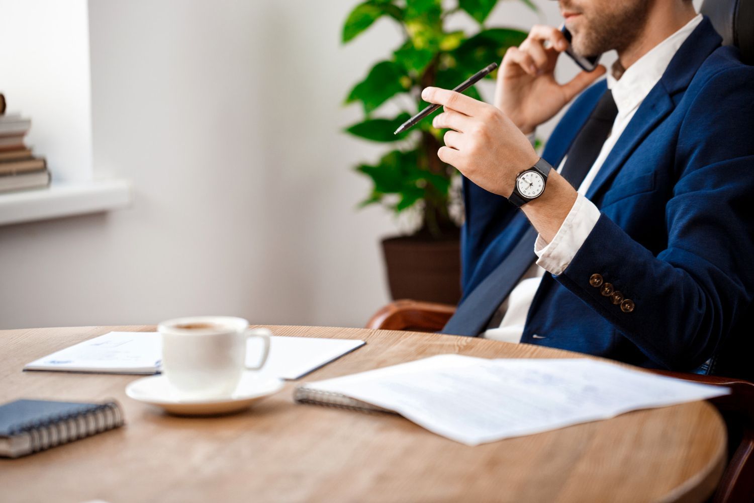 Businessman seated at a table, holding a phone beside a coffee cup and papers in a bright office setting