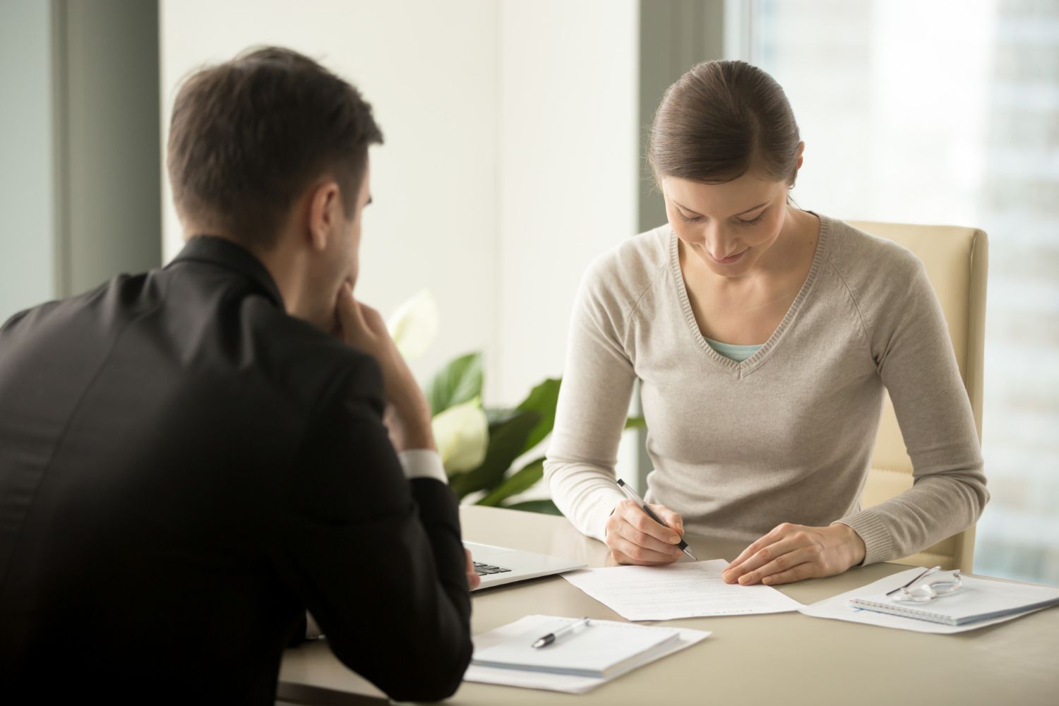 Two people in a meeting, one writing on paper at a table in a bright office
