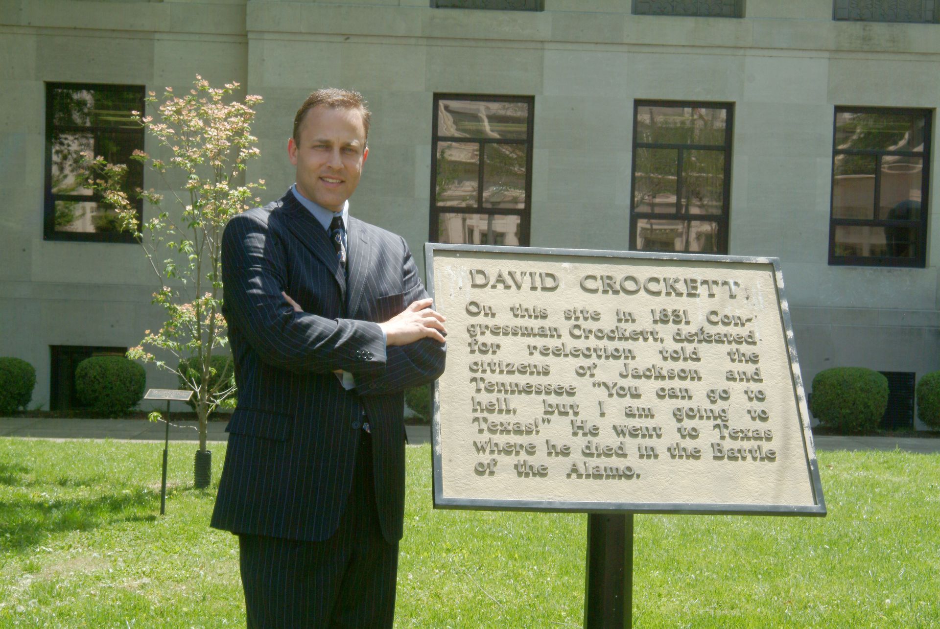 Man standing beside a historical marker sign on a lawn in front of a building