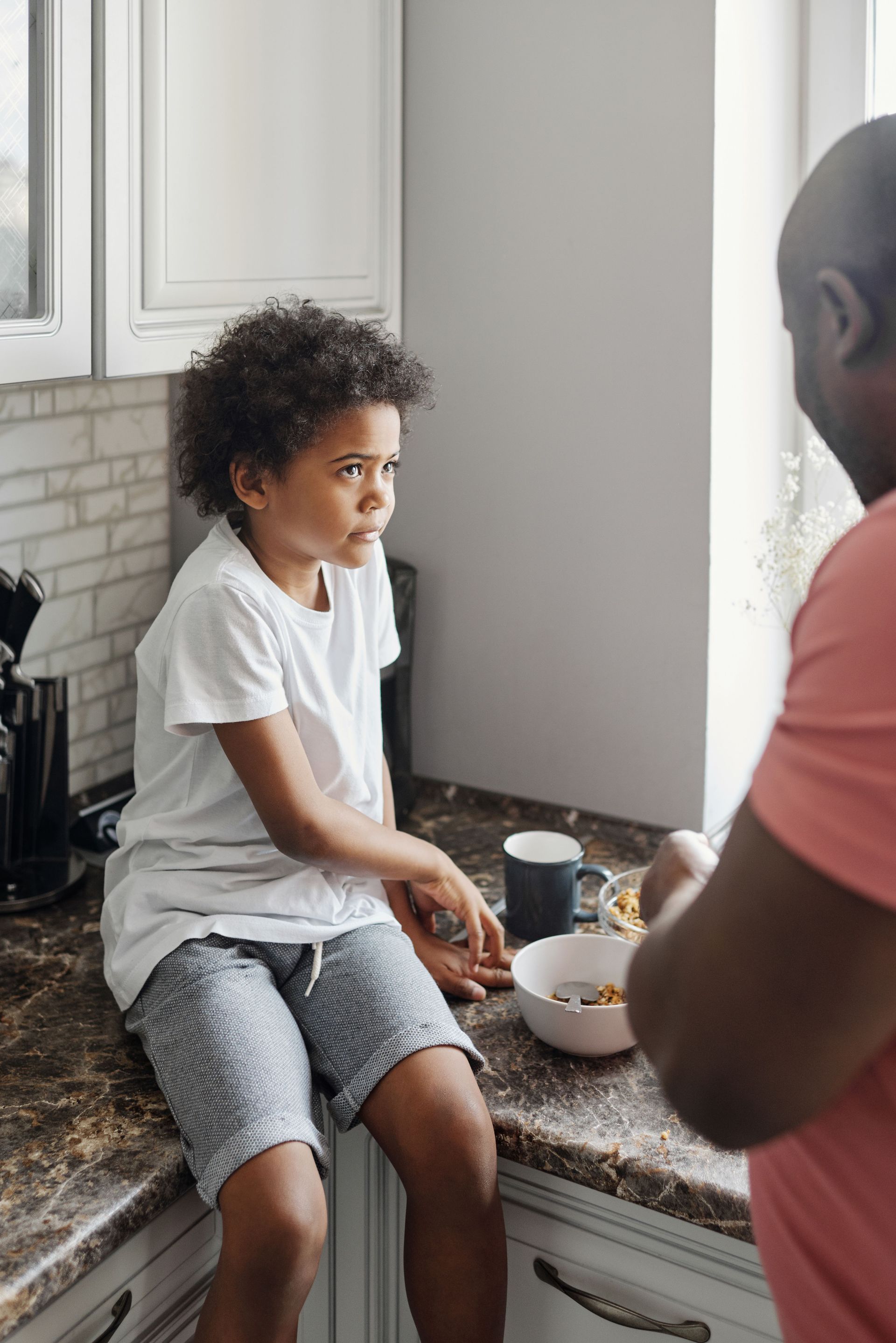 Child sitting on a kitchen counter, talking with an adult beside a bowl and cup.