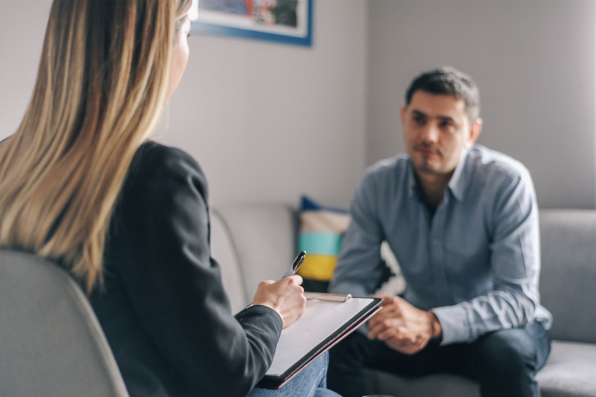 A woman is sitting on a couch talking to a man while holding a clipboard.