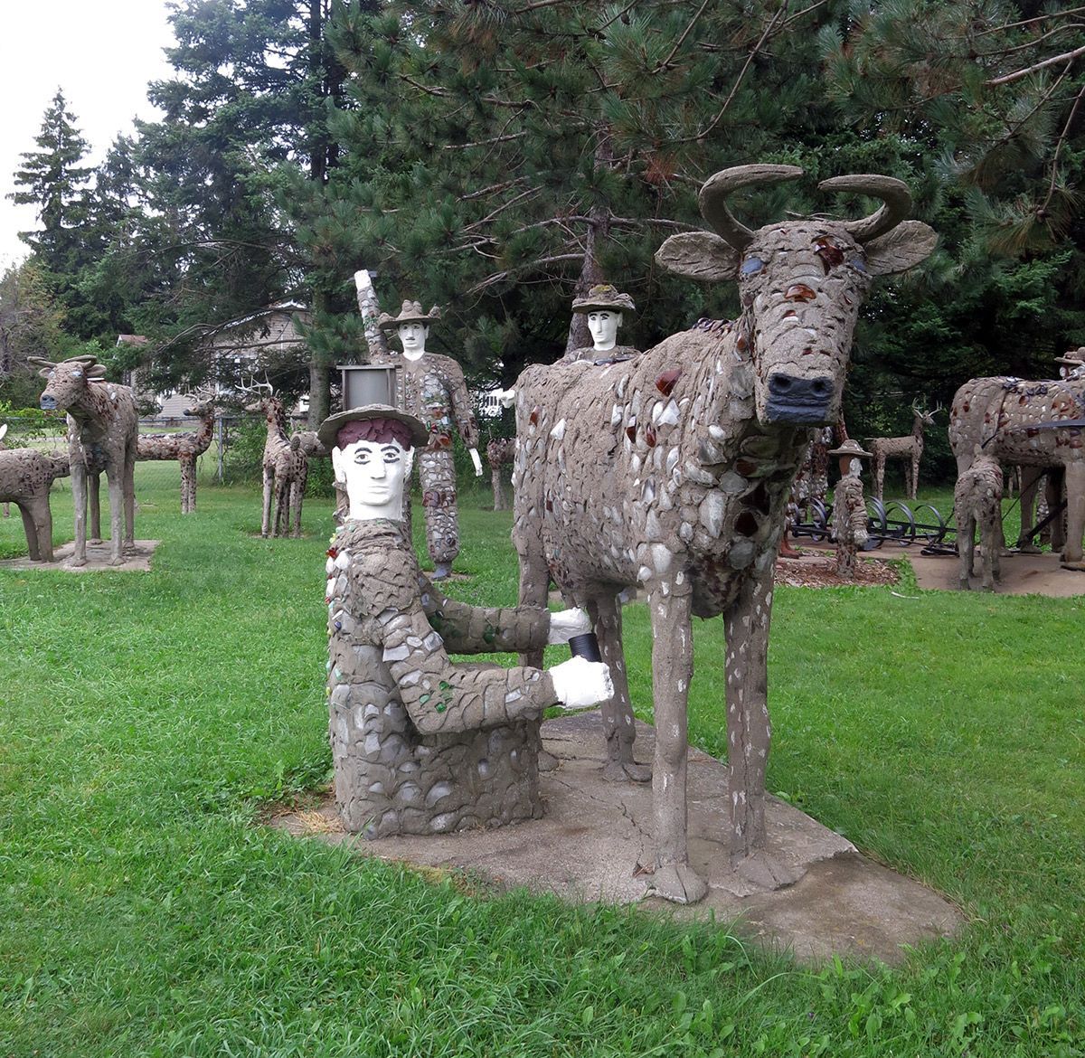 Stone sculptures of a seated person milking a cow in a grassy park with trees