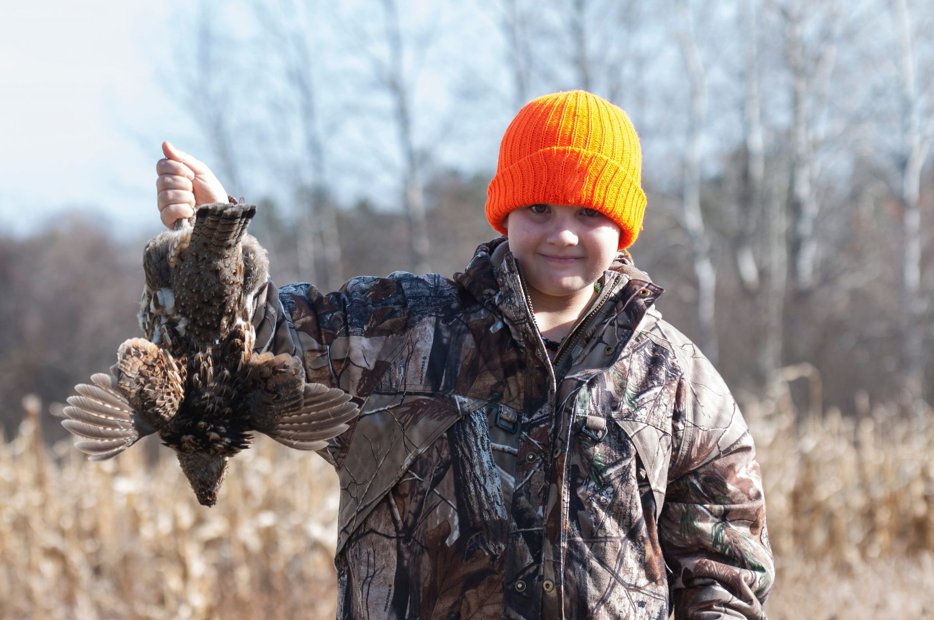 Person in camouflage and orange hat, holding a ruffed grouse outdoors.