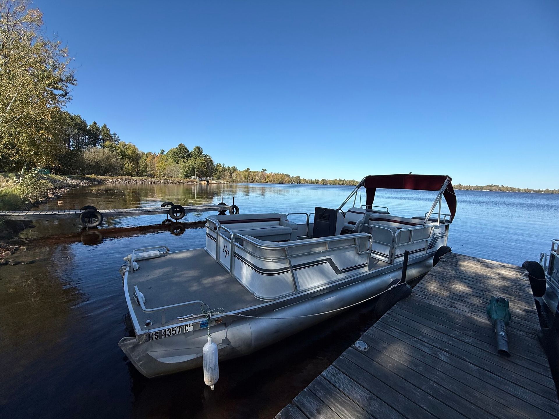 Pontoon boat docked on a wooden pier next to a lake under a clear, blue sky.