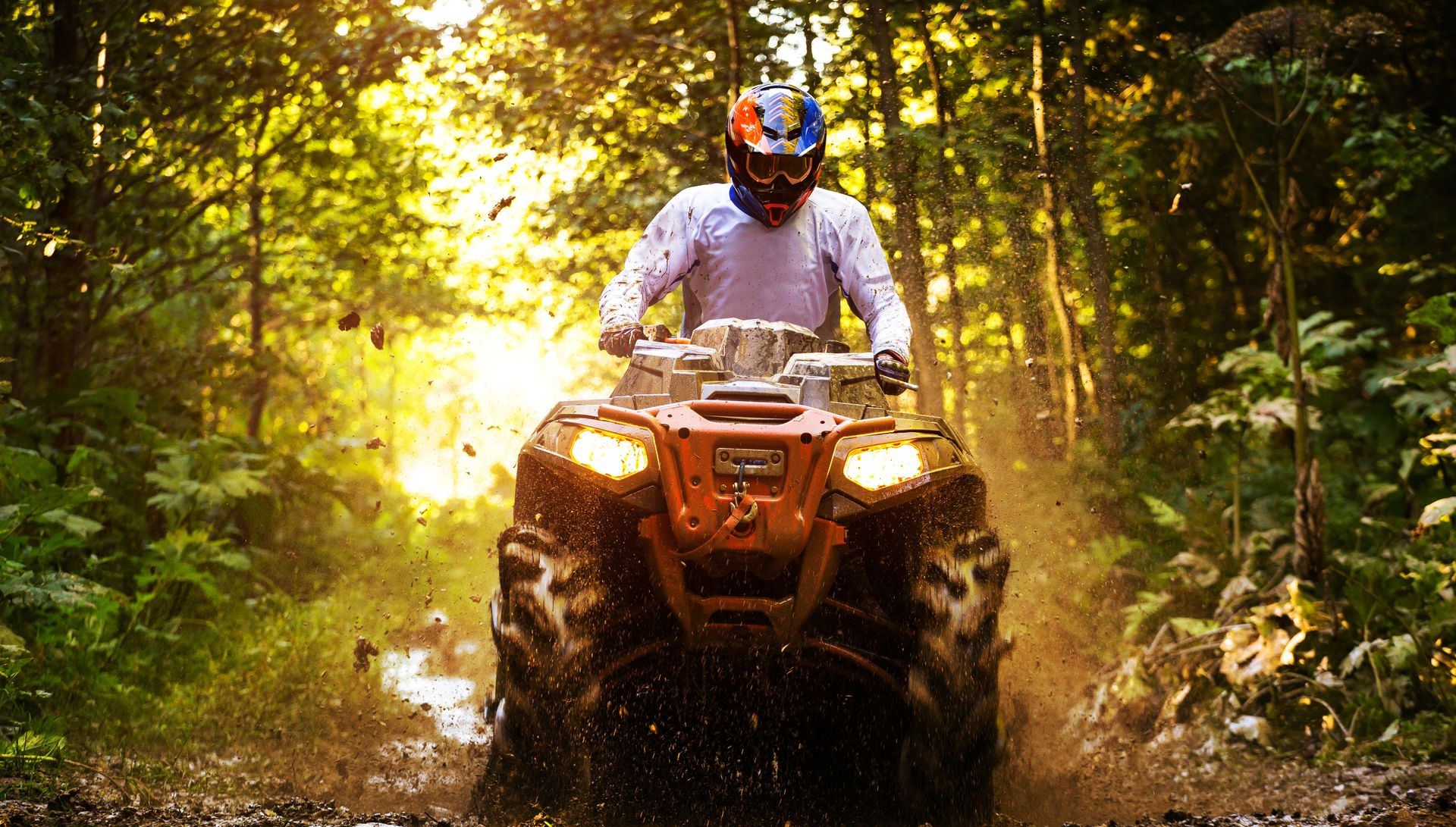 ATV rider in a helmet driving through a muddy forest trail with bright sunlight behind him