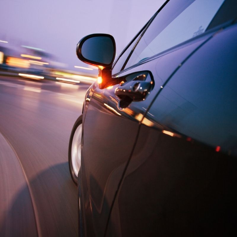 Close-up of a dark car speeding on a road at dusk, with motion blur and city lights streaking behind it
