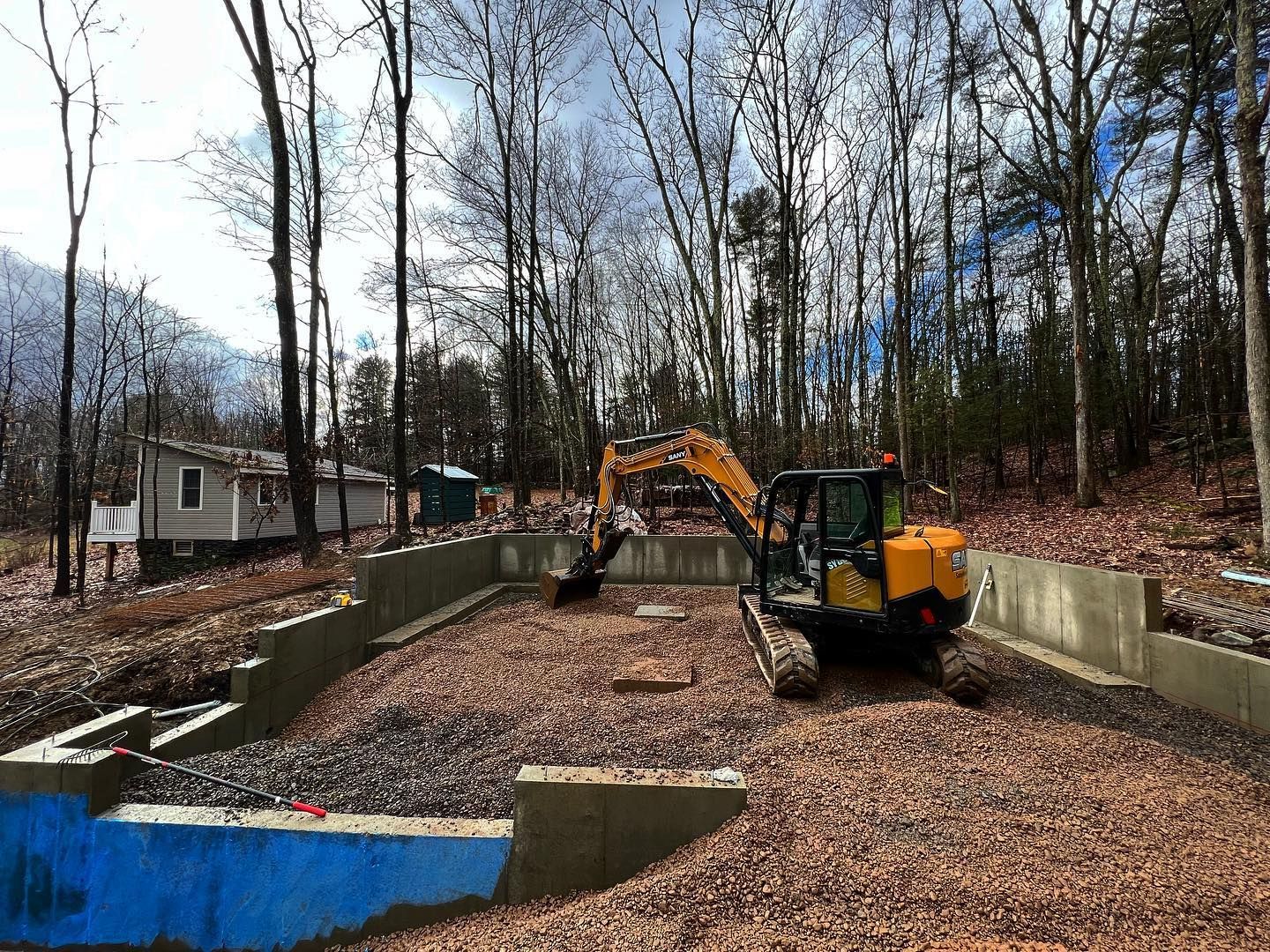 A yellow excavator is working on a swimming pool in the woods.