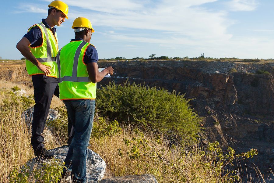 Two workers in hard hats and neon safety vests stand on a rocky edge overlooking a large quarry on a sunny day.