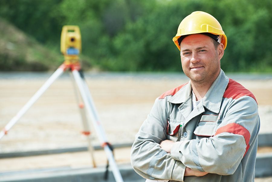 A construction professional wearing a yellow hard hat and work uniform, standing with arms crossed before a survey tripod.
