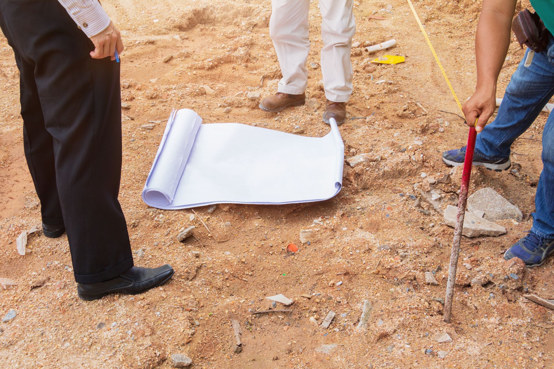 Three people stand on a dirt construction site looking down at a large rolled-out blueprint on the ground.