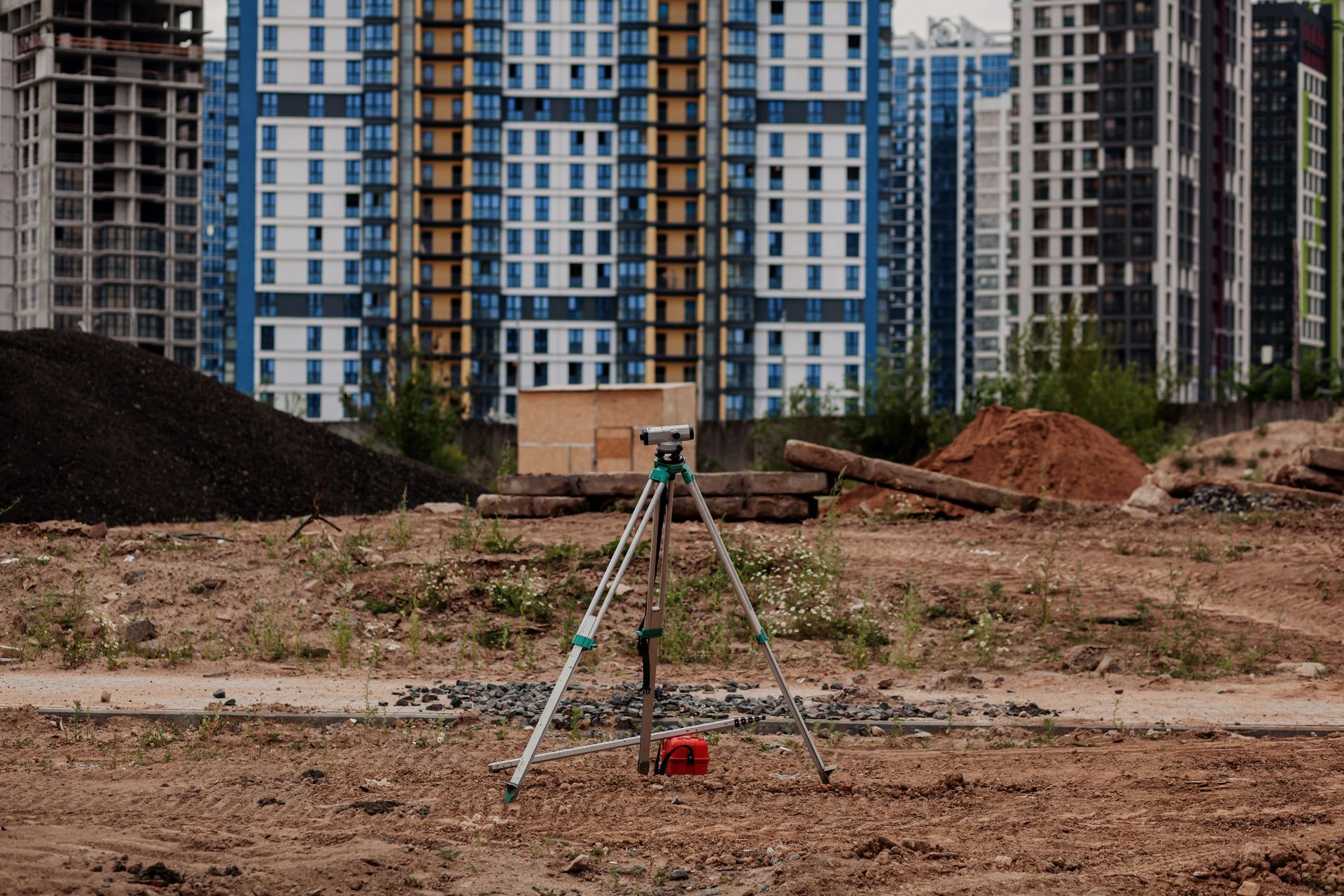 A surveying instrument on a tripod stands on a dirt construction site in front of multi-story residential buildings.