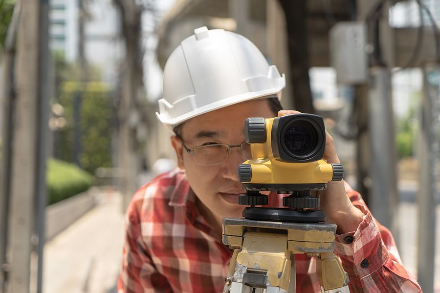 A surveyor in a white hard hat and plaid shirt looks through a yellow optical level on a tripod outdoors.