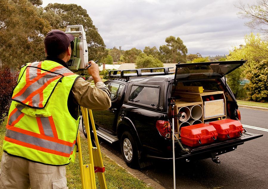 A land surveyor in a high-visibility vest uses a theodolite near a black truck parked on a residential street.