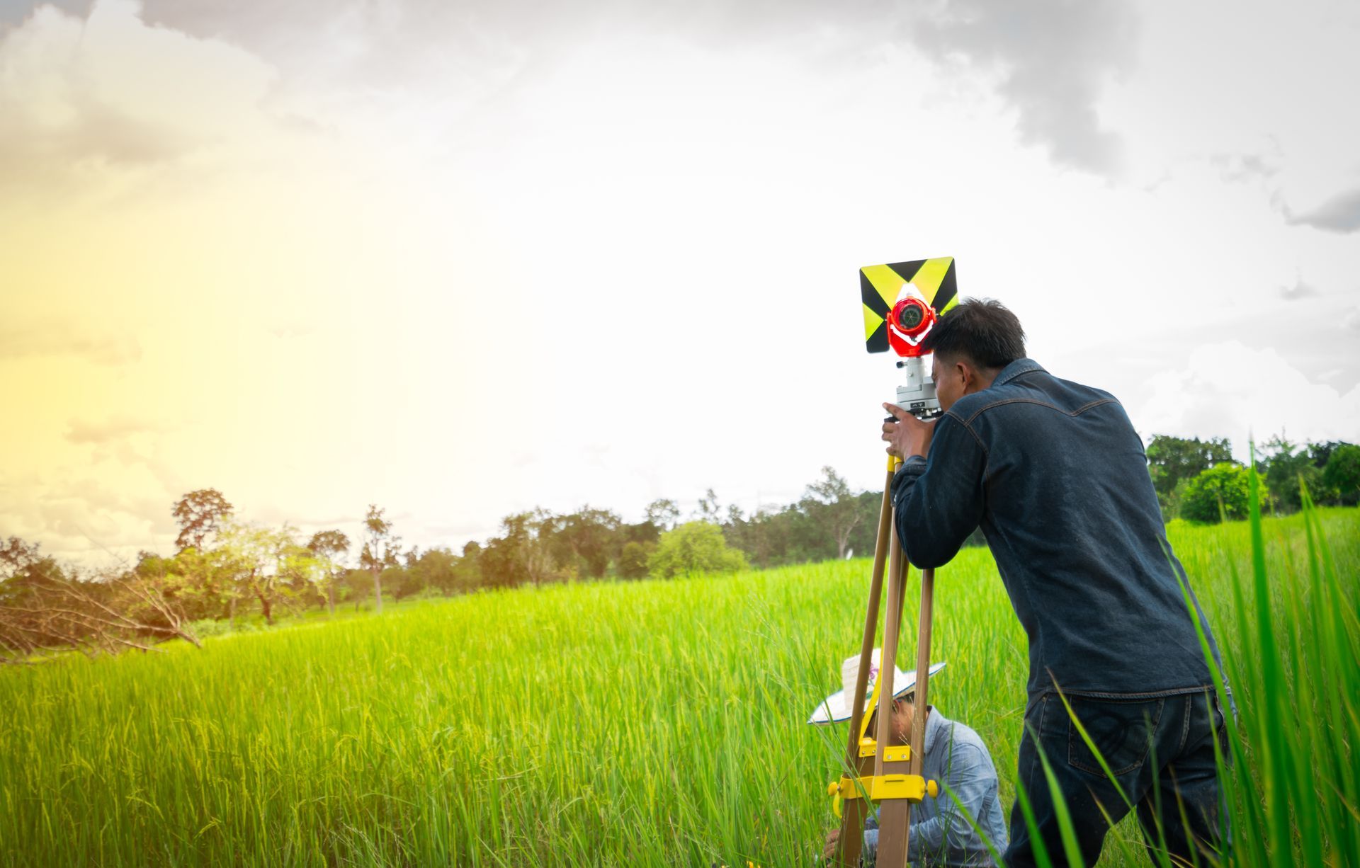 A surveyor uses a tripod-mounted prism target in a green field while another person works in the background.