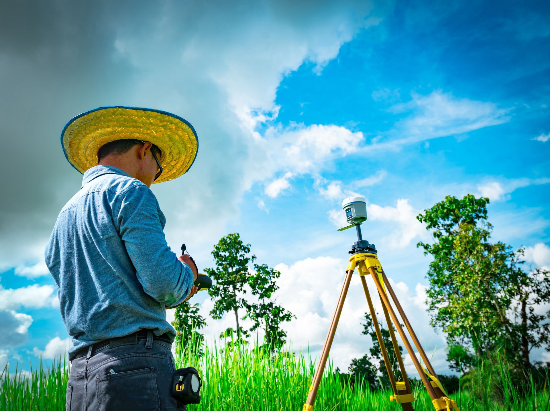 A person wearing a straw hat uses a handheld device to operate surveying equipment on a tripod in a grassy field.