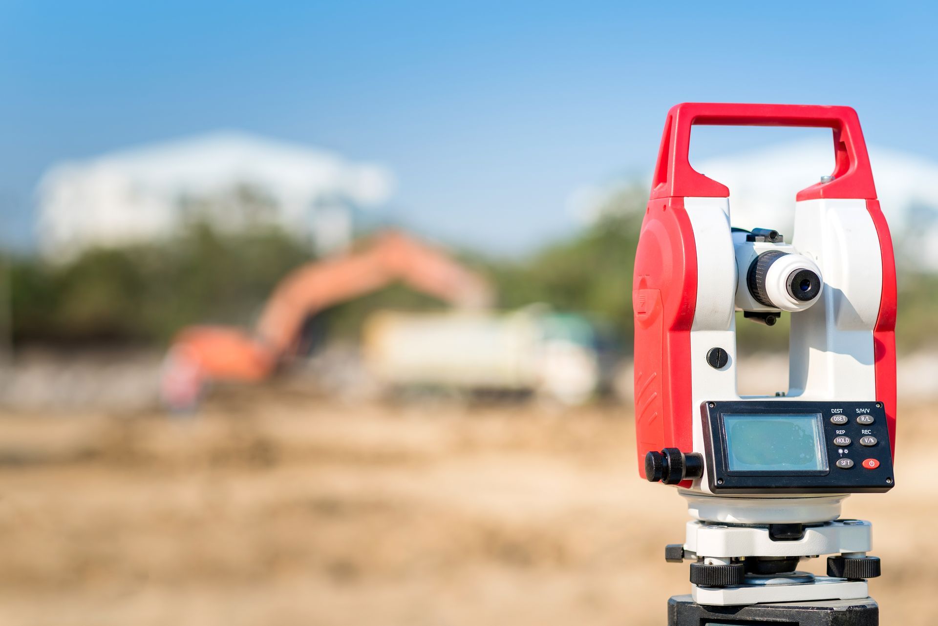 A red and white surveying total station on a tripod, with an out-of-focus construction site and excavator in the background.