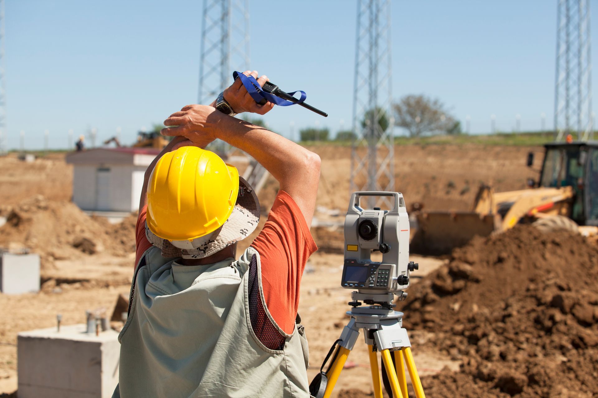 A surveyor in a yellow hard hat and vest uses a total station device at a construction site with heavy machinery nearby.