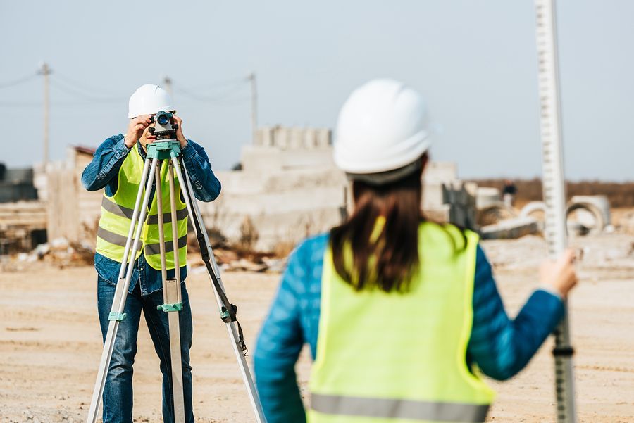 Two surveyors in high-visibility vests and hard hats work on a construction site, one using a tripod-mounted theodolite.