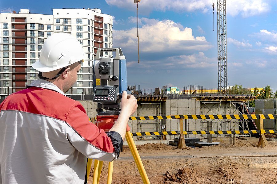 A surveyor in a white hard hat and red and white jacket uses a total station on a tripod at a construction site.