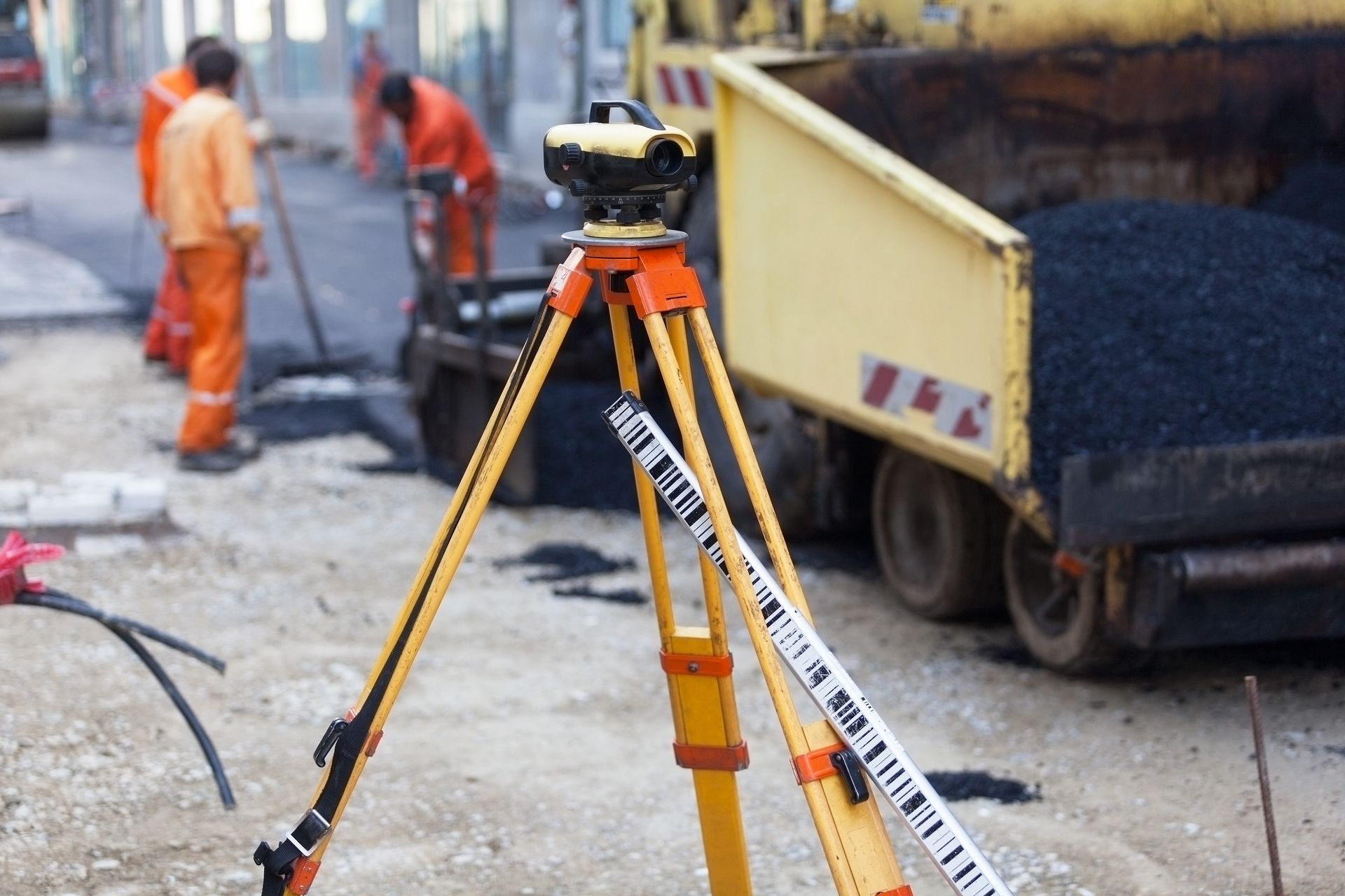 A survey level on a tripod in the foreground with construction workers and a truck paving a road in the background.