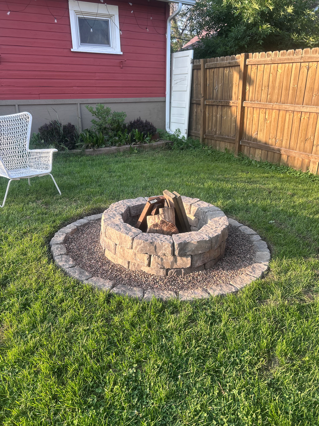 Backyard stone fire pit on a lawn beside a red house and wooden fence