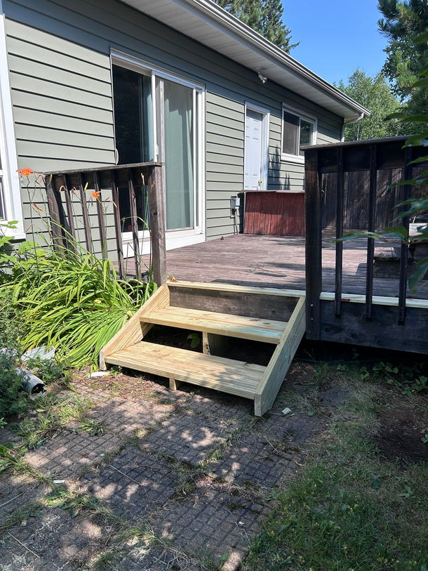 Wooden porch ramp leading to a small deck beside a gray house, with black railing and a brick landing.