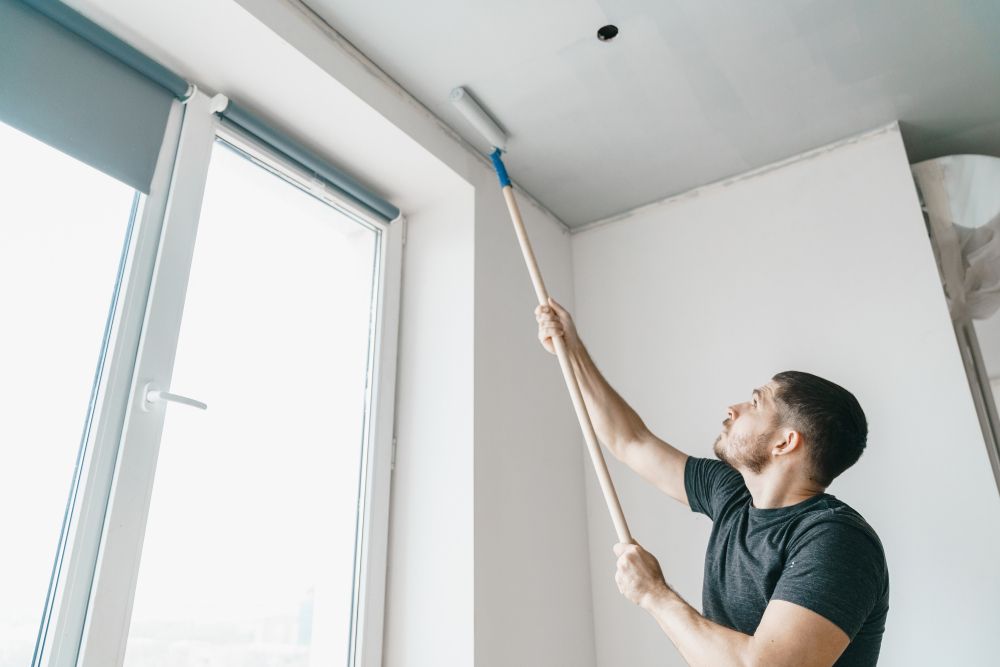 A Man Is Painting The Ceiling Of A Room With A Roller — Auslinc Building Services In Wingham, NSW