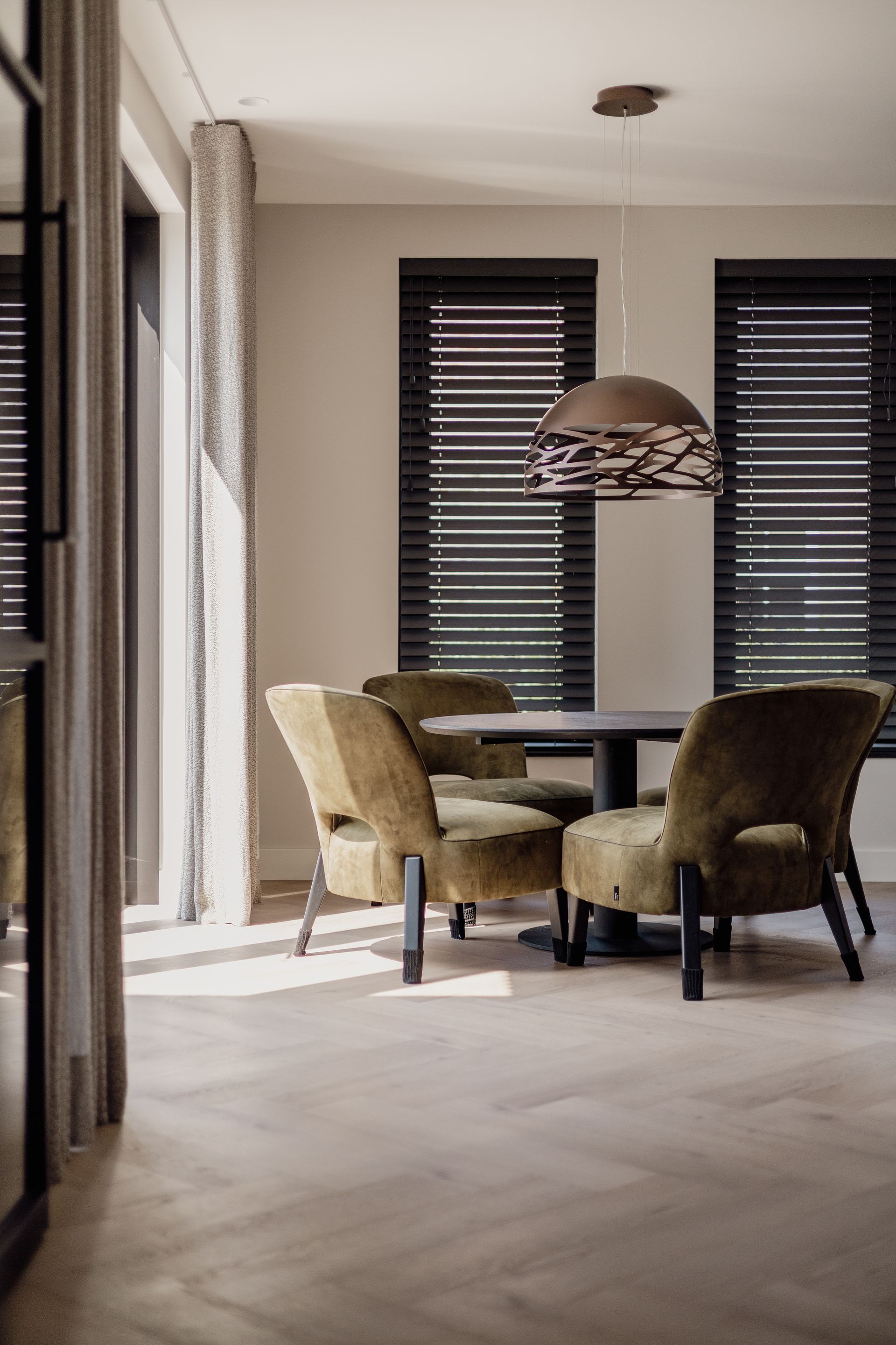 Moderne eetkamer met een ronde tafel, fluwelen stoelen, hanglamp en donkere raamjaloezieën.