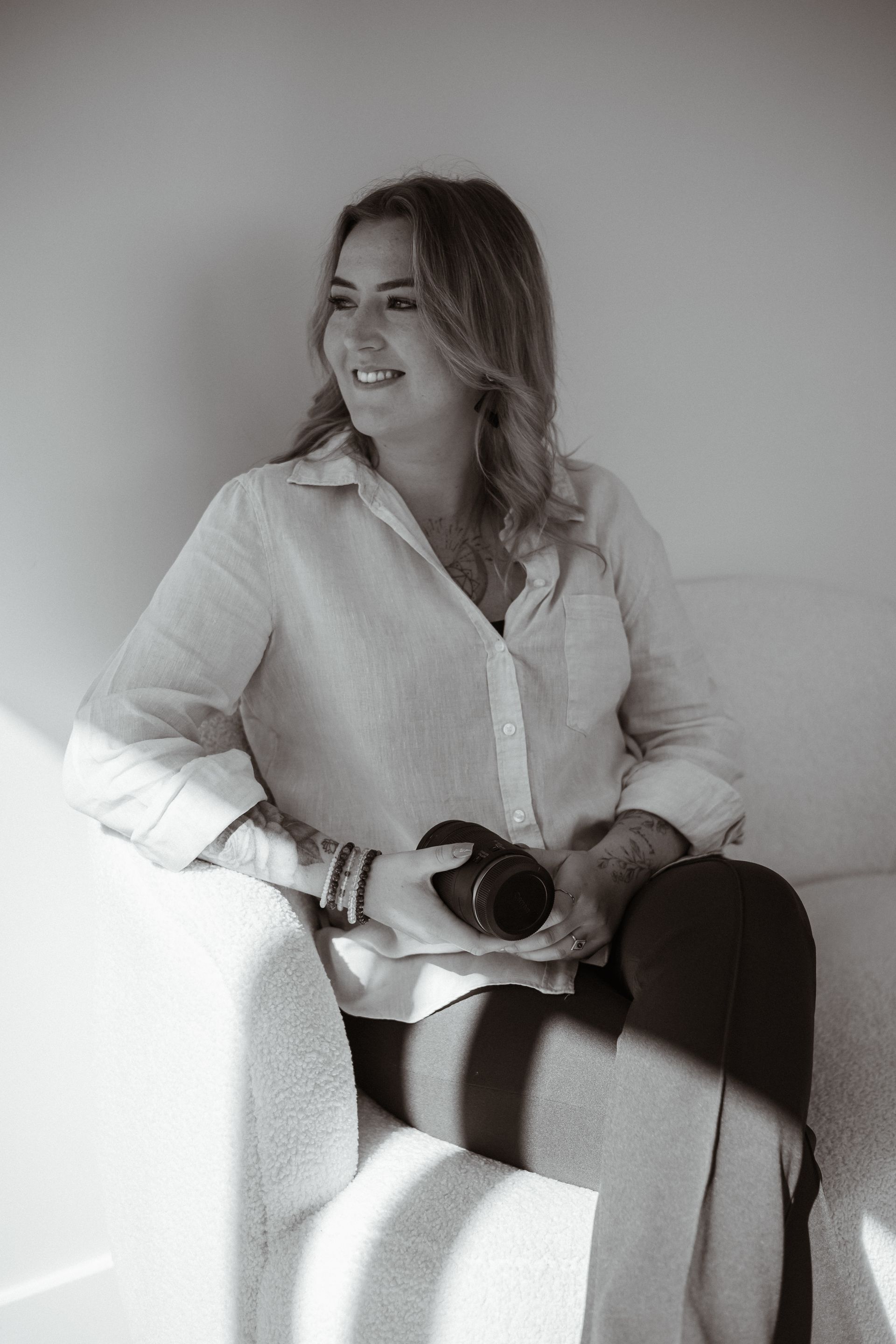 Vrouw in een lichte blouse zit bij een raam, glimlachend en met een lens in haar hand.