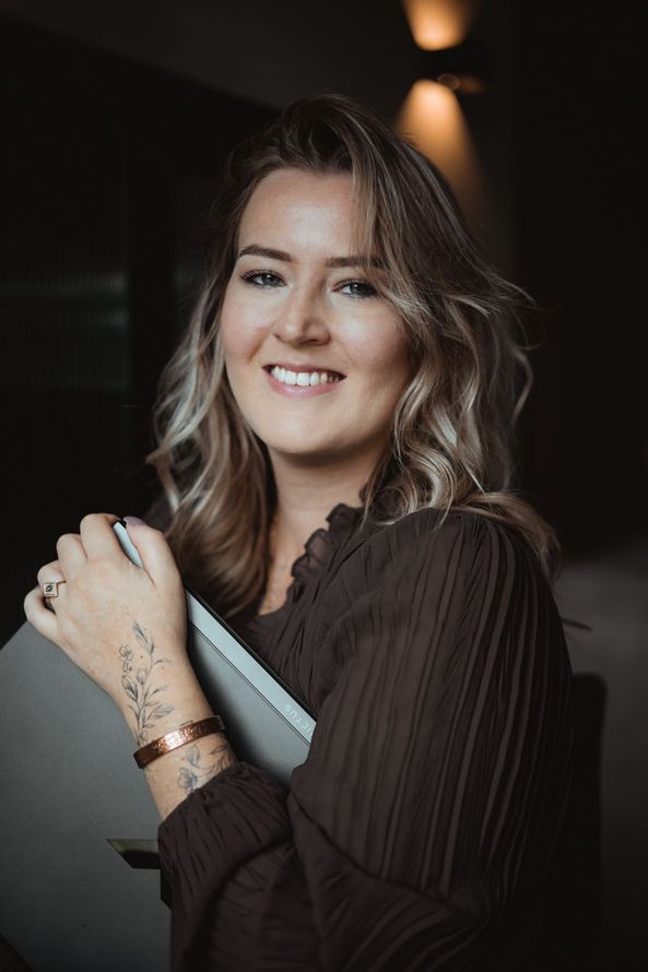 Vrouw in een lichte blouse zit bij een raam, glimlachend en met een lens in haar hand.