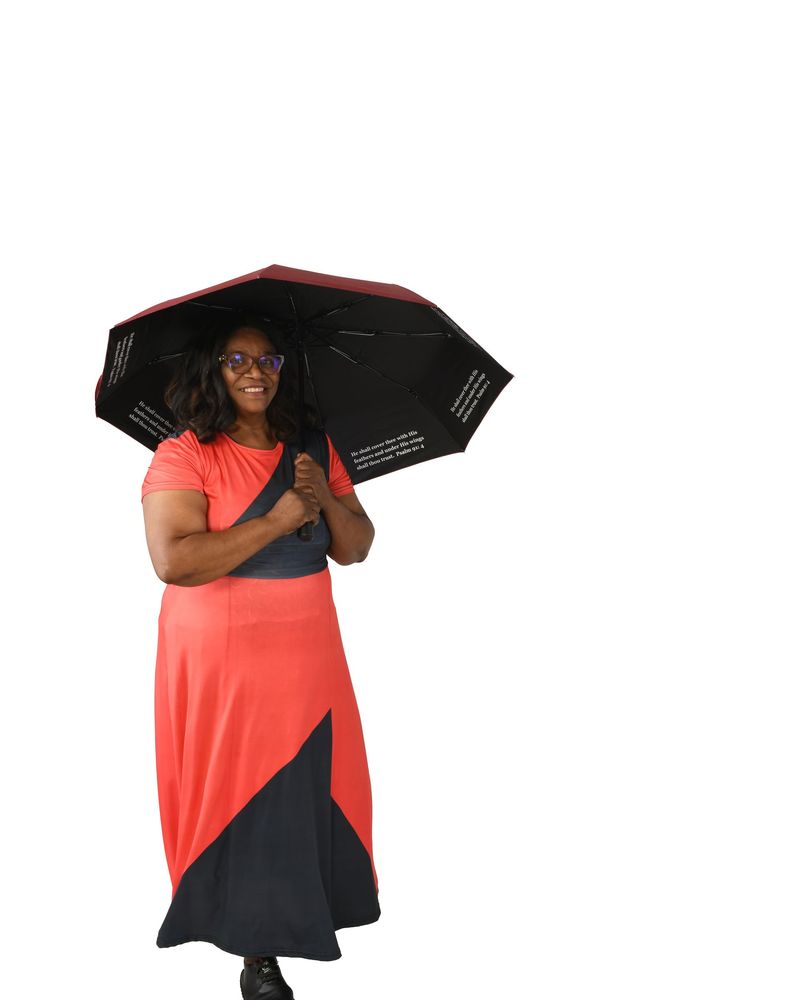 Person jumping over colorful umbrellas while holding a gray umbrella in a grassy field under a blue sky