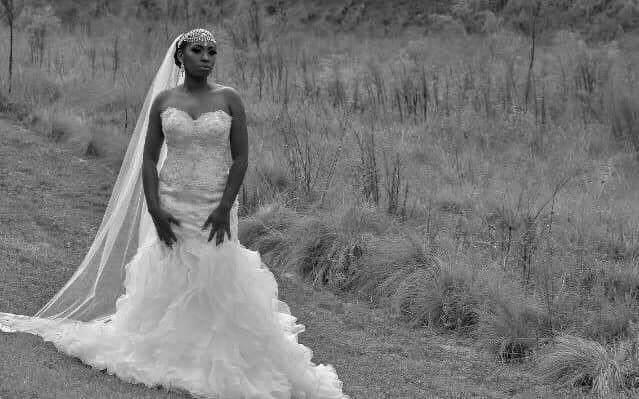A black and white photo of a bride in a wedding dress and veil standing in a field.