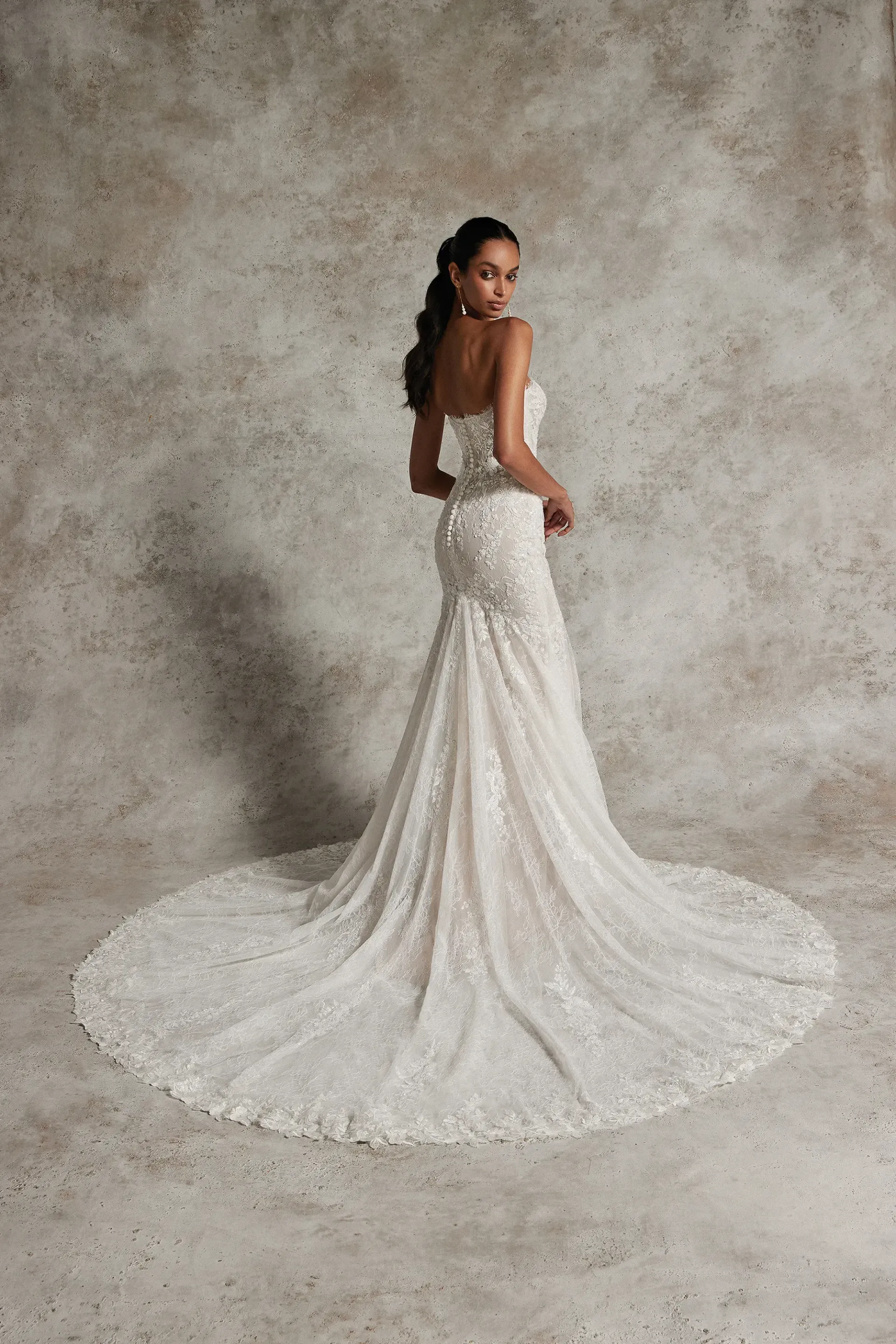 A model in a strapless, floor-length white lace mermaid wedding dress with a long train, standing against a neutral wall.