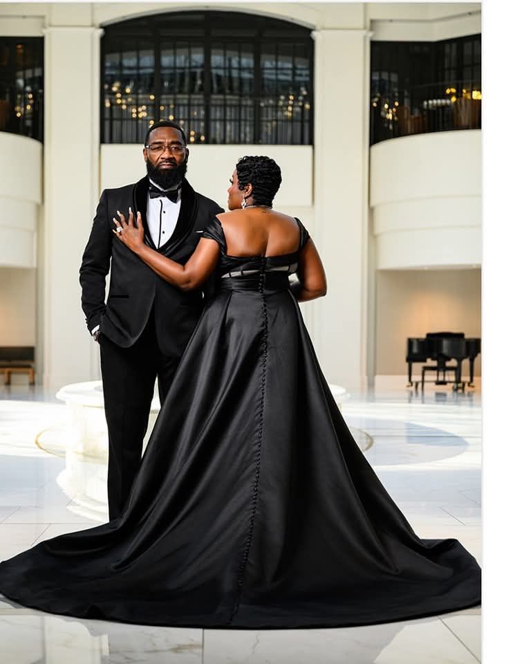 A couple dressed in formal black attire stands in an elegant, sunlit atrium with a grand staircase and a piano.