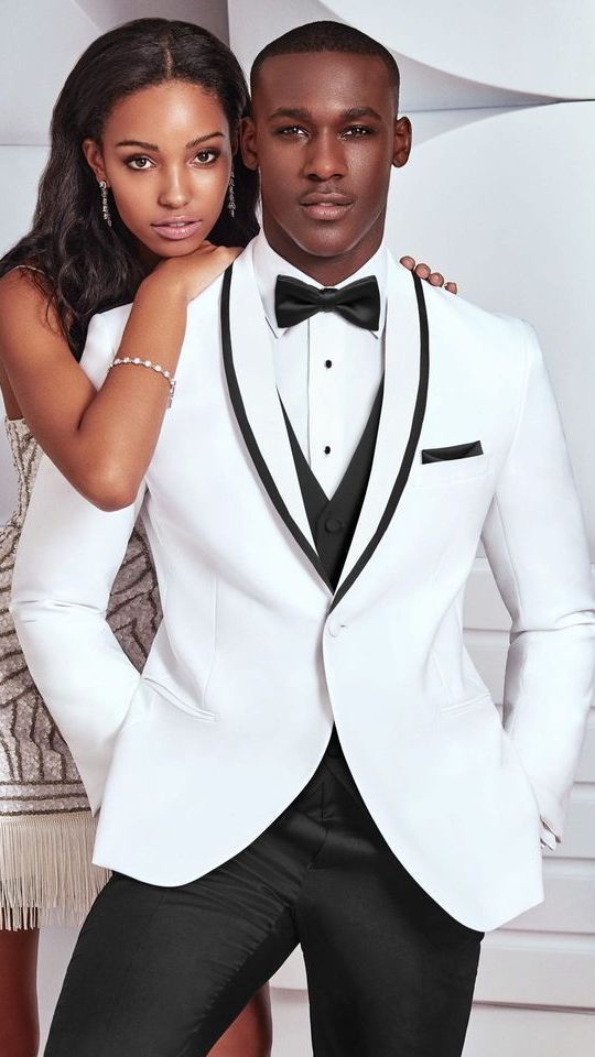 A man in a white tuxedo with a black shawl lapel and bow tie stands with a person wearing a beaded dress.
