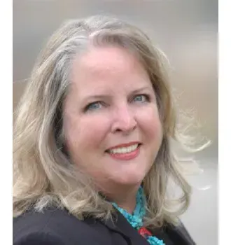 Woman with graying blonde hair smiling, wearing a turquoise necklace and black blazer.
