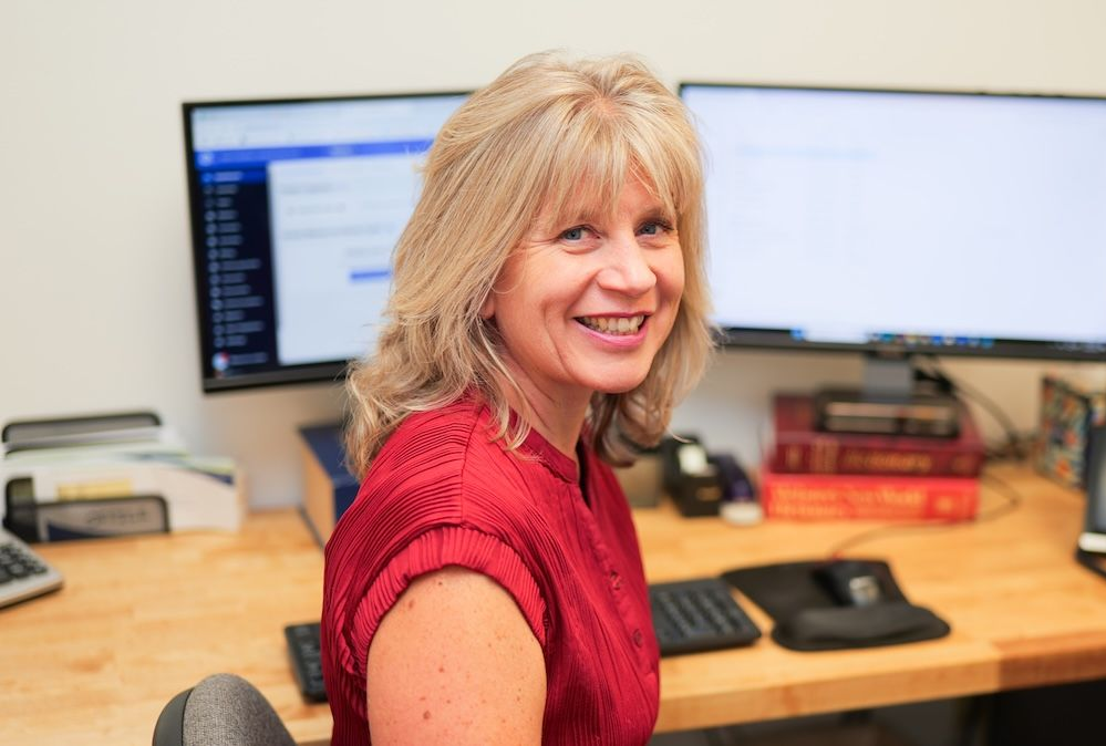 Woman smiling at desk with two computer monitors, wearing a red sweater.