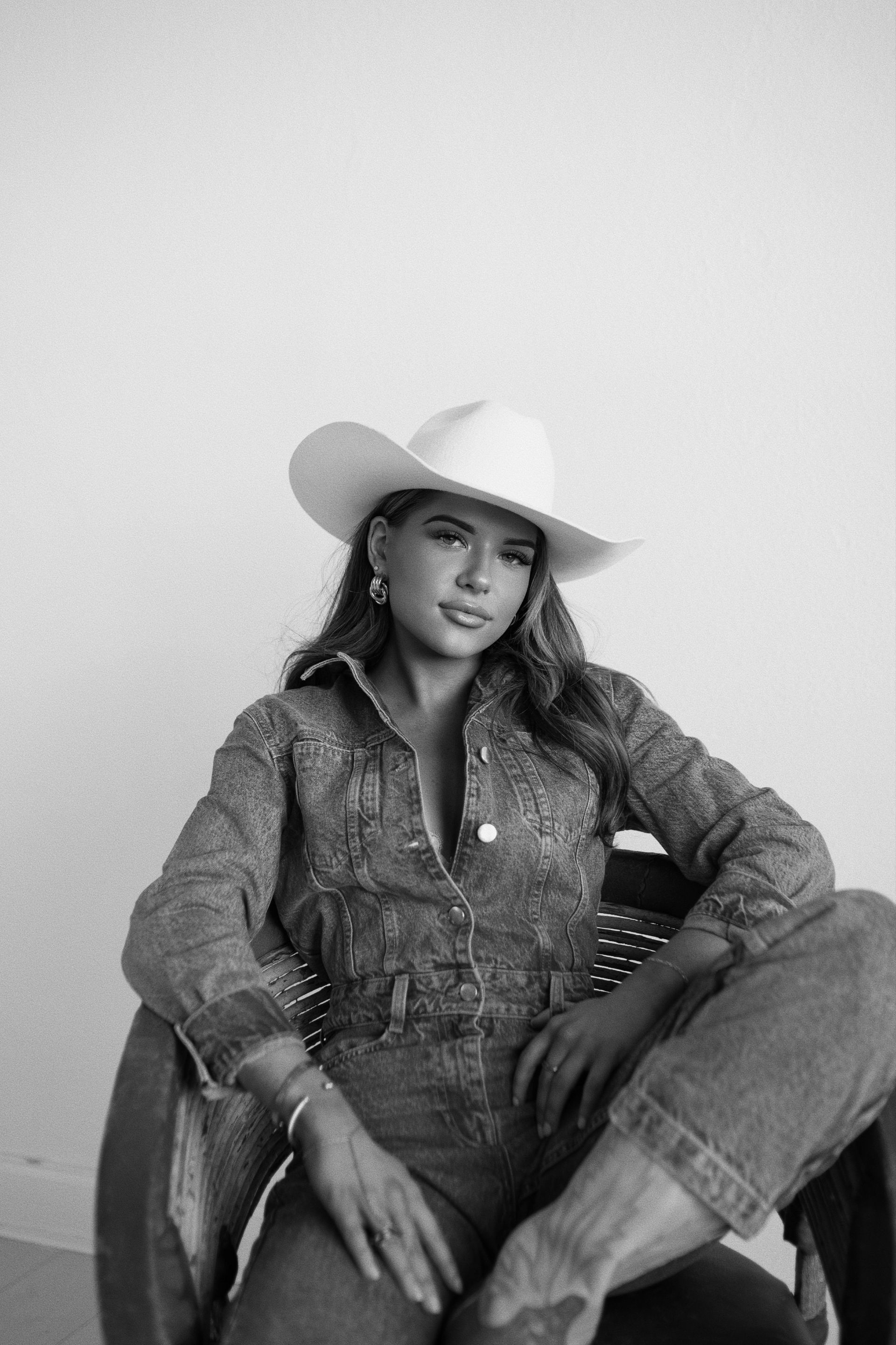 Ava in a cowboy hat seated on a chair, wearing denim and looking at the camera.