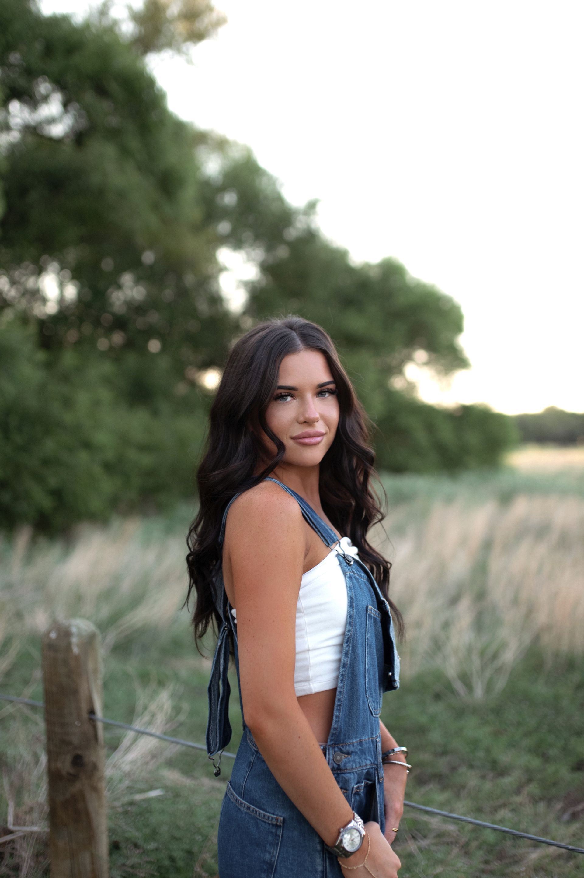 Ava standing outdoors in overalls, looking at the camera near a grassy field and trees.